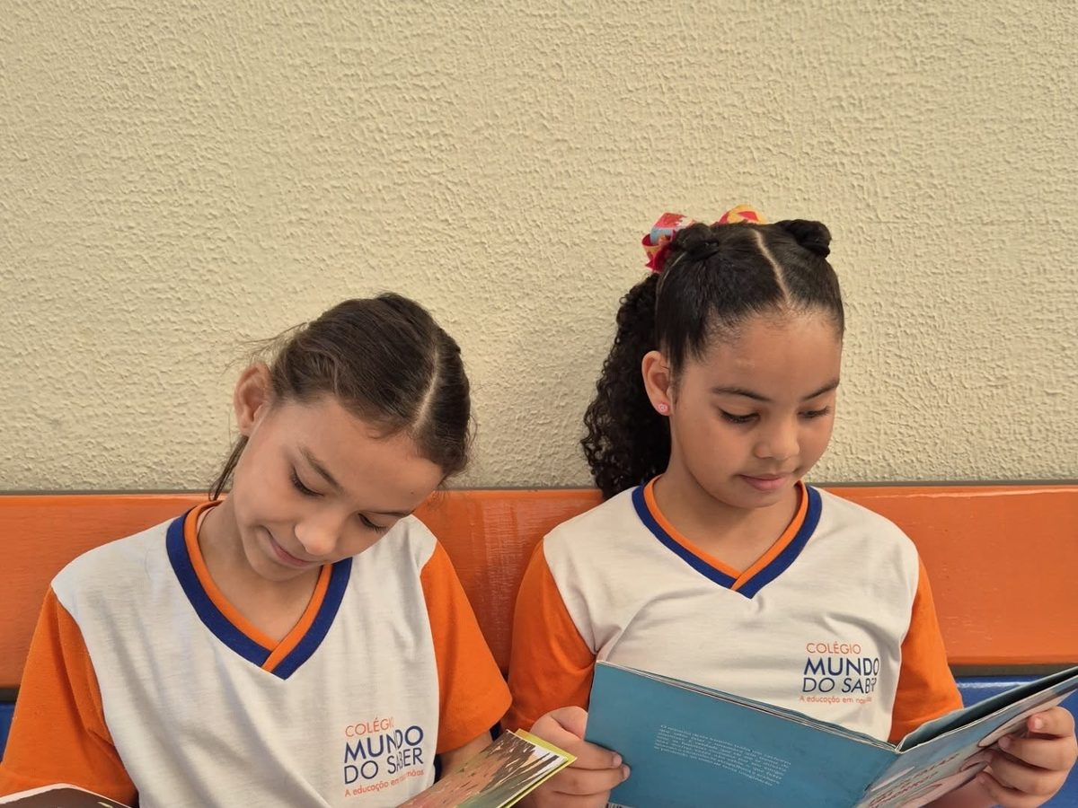 Duas alunas de uniforme lendo o mesmo livro, sentadas lado a lado